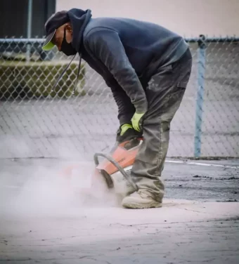 A construction worker in safety gear cuts concrete on an outdoor pavement.