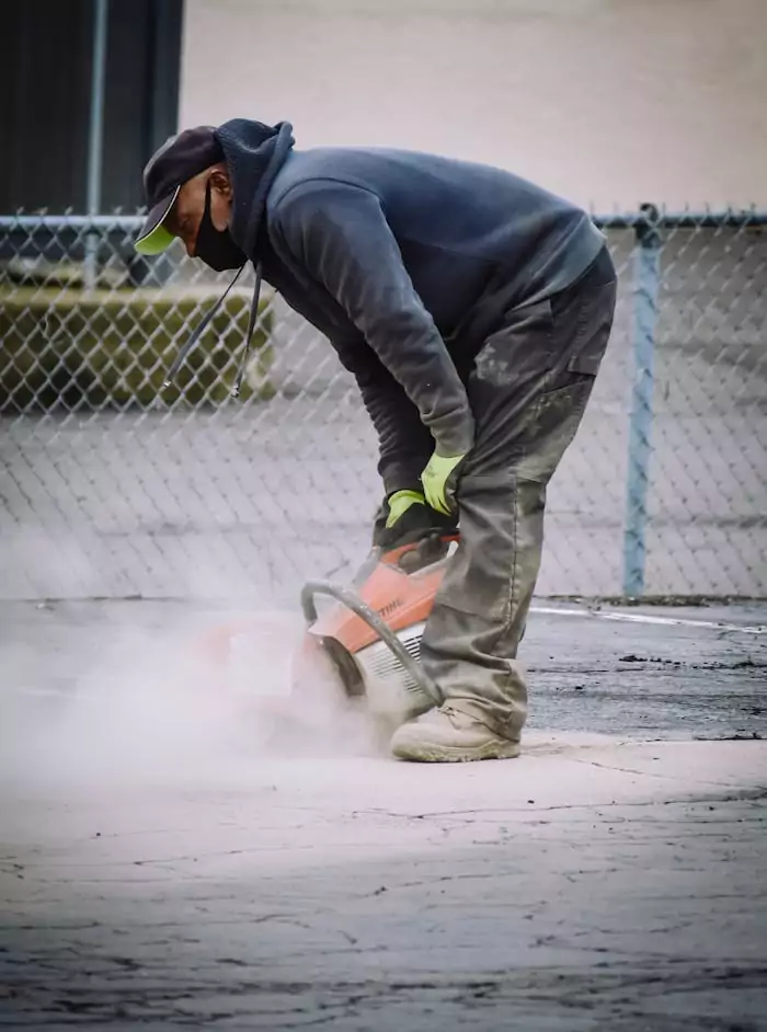 A construction worker in safety gear cuts concrete on an outdoor pavement.